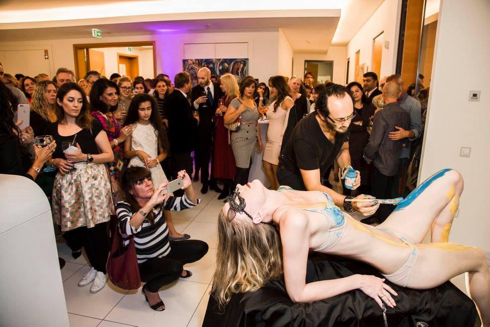 Body painting a reclining model at an indoor event, guests watching and filming