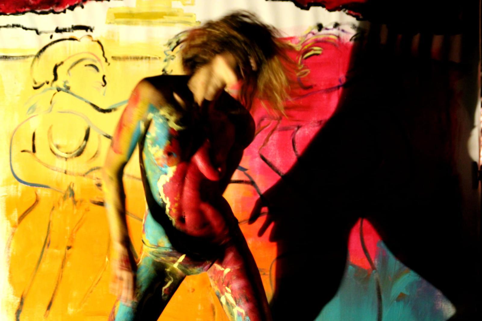A body-painted dancer moving in front of the canvas, lit against the dark